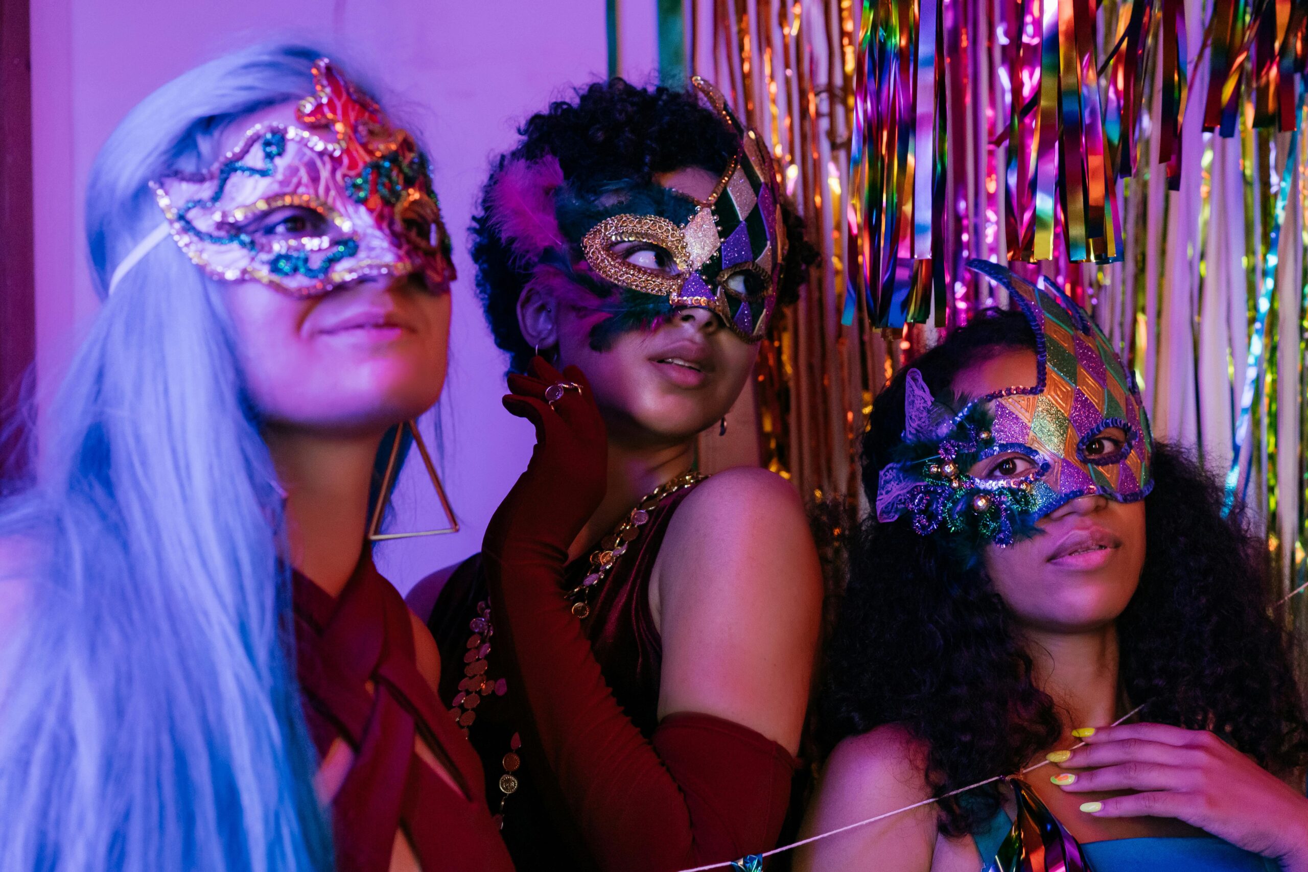 Three women in masquerade masks celebrate at a vibrant party with colorful decorations.